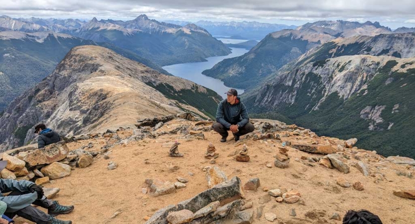 A person sits on a rocky overlook. Behind them is a vast mountain landscape with a river winding through it. 